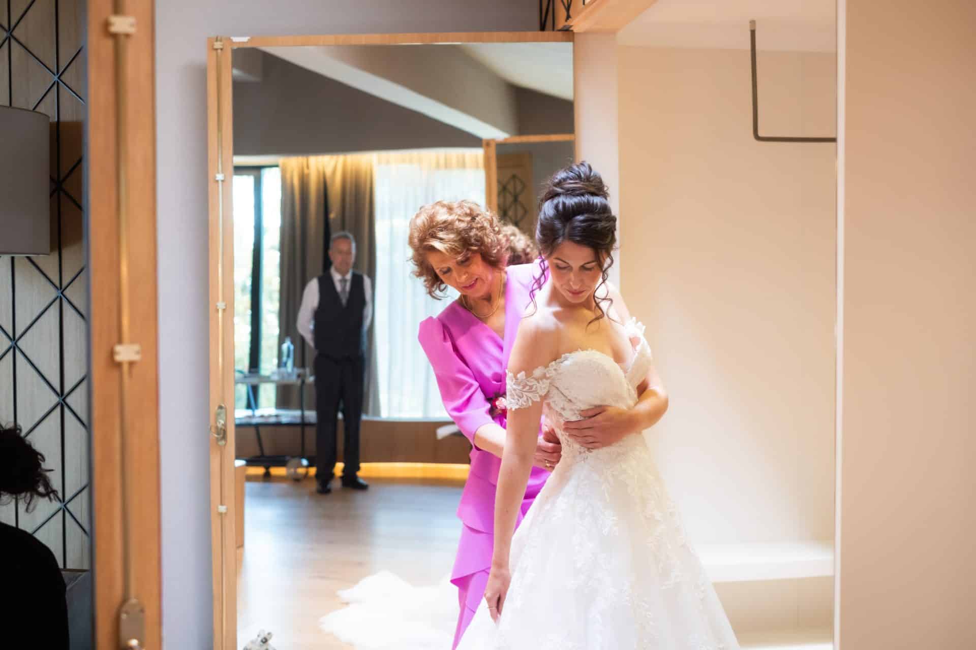 La madre ayudando a Diana a colocarse el vestido de novia mientras el padre espera antes de la ceremonia
