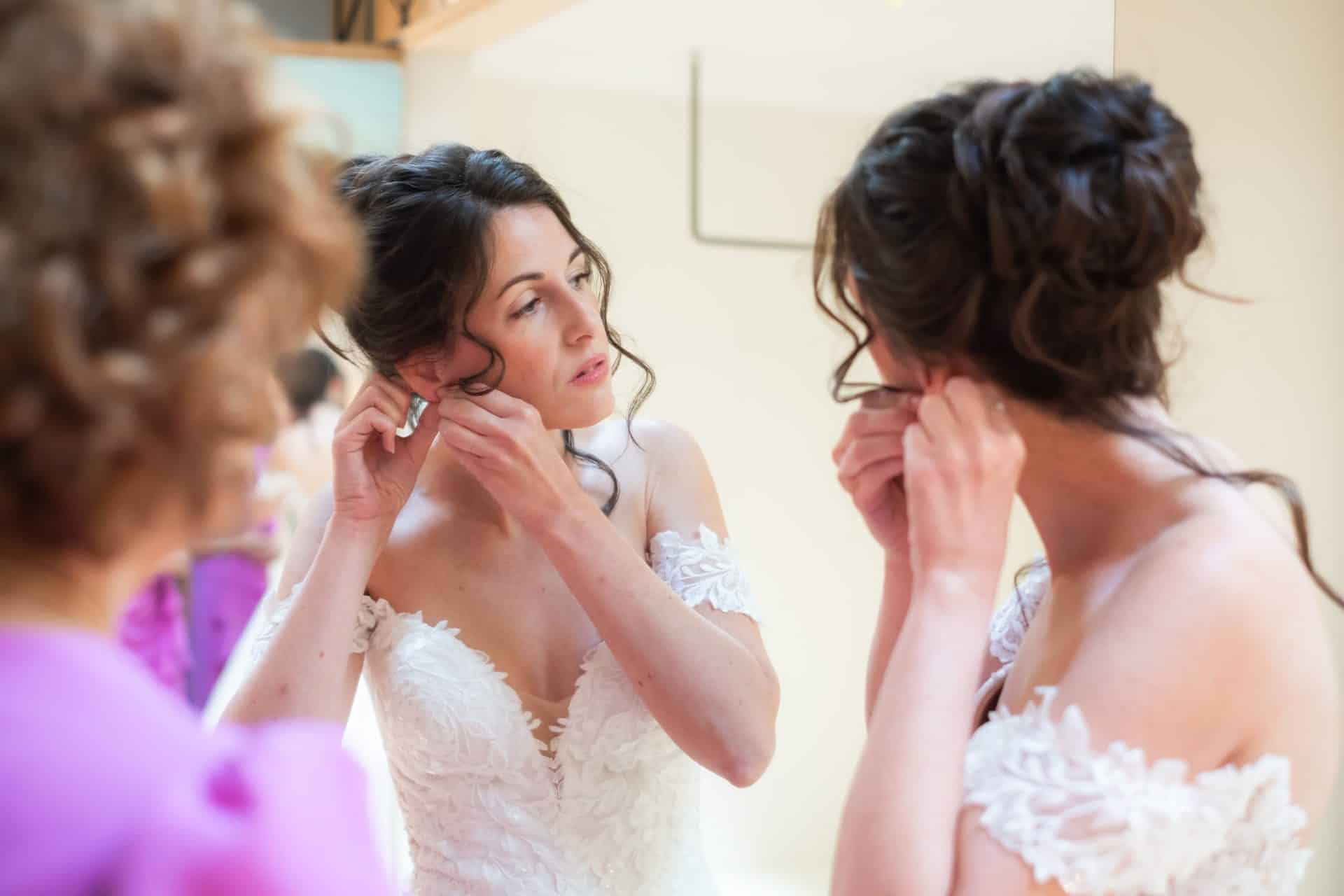 Diana colocándose un pendiente frente al espejo antes de la ceremonia con maquillaje natural y recogido de novia